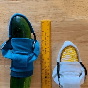May include: A cucumber and a corn cob are each wearing a blue and white hoodie, respectively. The text "One Size Fits All!" is at the top of the image. A yellow ruler is to the right of the cucumber.