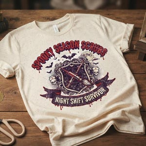 May include: A cream-coloured t-shirt featuring a graphic print with the text "SPOOKY SEASON SCRUBS" and "NIGHT SHIFT SURVIVOR." The design includes a central shield with medical instruments, skulls, bats, and a moon, all rendered in red, purple, and black.