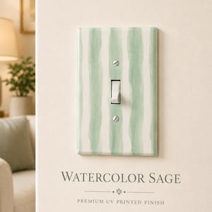 May include: A rectangular light switch wall plate featuring a vertical sage green watercolor stripe pattern on a white background. The plate includes a central white toggle switch and two silver screws for mounting, with the text WATERCOLOR SAGE.
