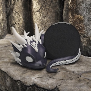 Puede incluir: Un altavoz con forma de dragón negro y blanco con una rejilla de altavoz redonda negra. El dragón está enroscado con la cabeza apoyada en el altavoz.