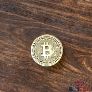 May include: A gold-colored Bitcoin coin with the Bitcoin symbol in the center. The coin has intricate details and text around the edges. The coin is on a dark wood surface.
