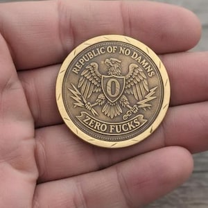 Puede incluir: Una moneda redonda de color oro antiguo con el texto "REPUBLIC OF NO DAMNS" y "ZERO FUCKS". La moneda presenta un águila con un escudo y el número "0". La moneda está sujeta en una mano.