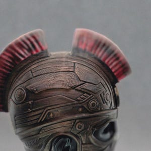May include: A bronze-colored helmet with a textured surface and a red and black crest. The helmet has a futuristic design with geometric shapes.