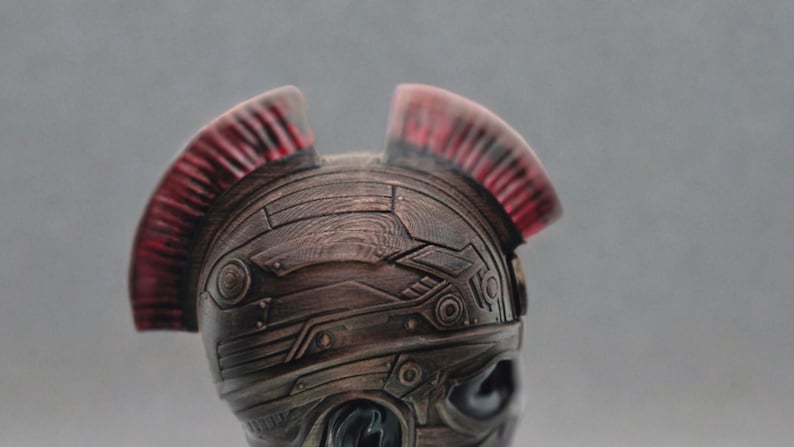 May include: A bronze-colored helmet with a textured surface and a red and black crest. The helmet has a futuristic design with geometric shapes.