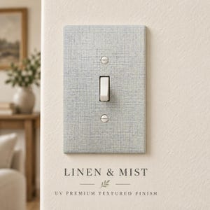 May include: A light gray light switch cover with a textured linen pattern, featuring a central white toggle switch and two silver screws. The text LINEN & MIST UV PREMIUM TEXTURED FINISH is printed below the switch plate on the wall.