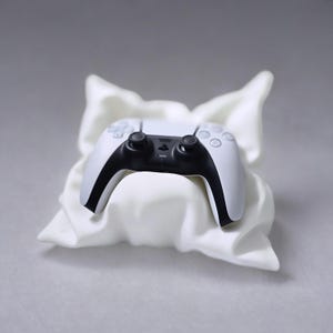 May include: A white and black video game controller resting on a white pillow-shaped stand.
