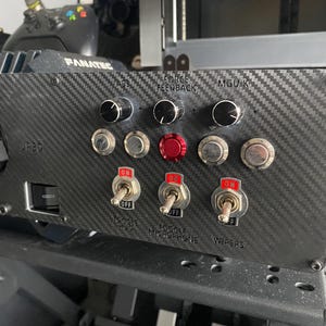 May include: A black carbon fibre panel with various switches and knobs. The panel has labels for "Force Feedback", "MGUK", "Toggle Microphone", and "Wipers".