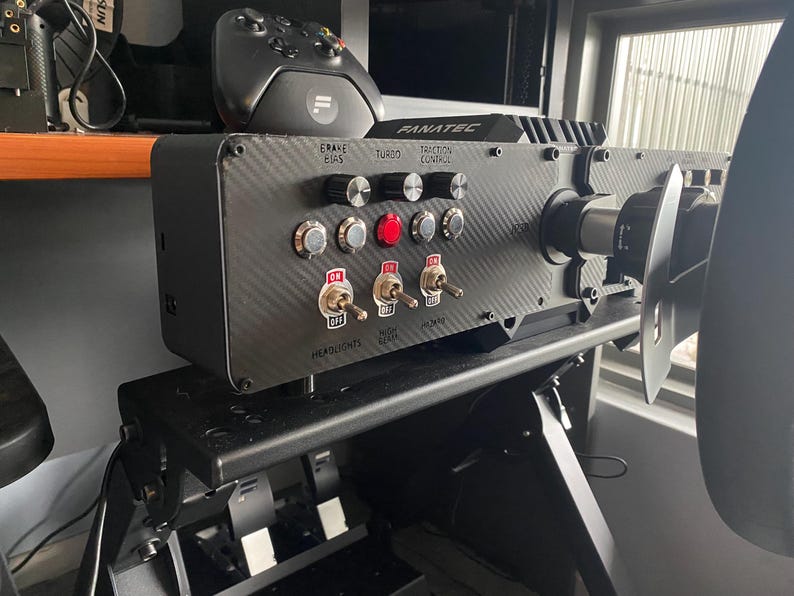 May include: A black racing wheel with a carbon fibre panel featuring buttons and switches labelled "Brake/Gas", "Turbo", "Traction Control", "Headlights", "Radio", "Seat", and "On/Off".