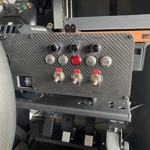 May include: A black carbon fibre panel with a variety of switches and knobs. The panel has labels for "Force Feedback", "MGU-X", "Toggle Push", "Toggle Microphone", and "Wipers".