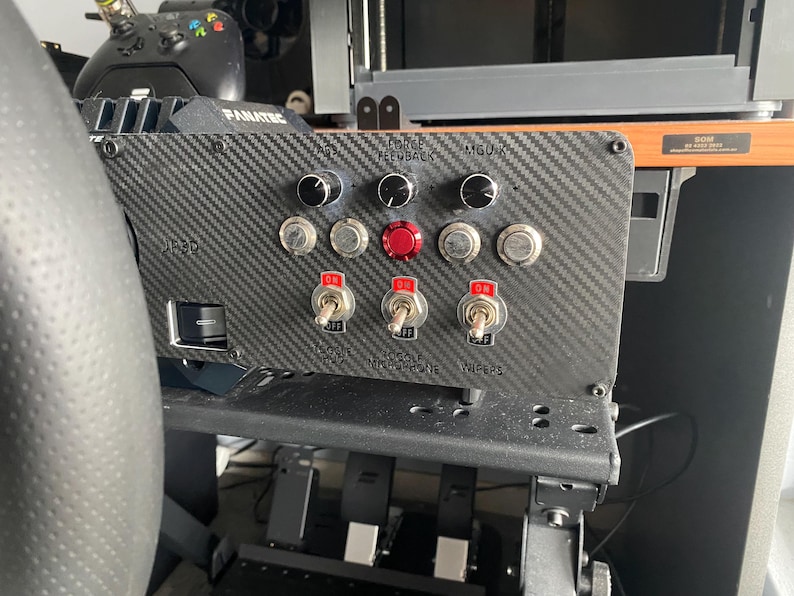 May include: A black carbon fibre panel with a variety of switches and knobs. The panel has labels for "Force Feedback", "MGU-X", "Toggle Push", "Toggle Microphone", and "Wipers".