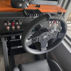 May include: A black racing wheel with a carbon fibre design and a black and silver base. The wheel has a variety of buttons and switches, including a red button on the right side. The wheel is attached to a black racing simulator rig.