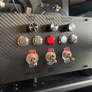 May include: A black panel with a carbon fibre texture, featuring various controls for a racing simulator. The controls include knobs for brake bias, turbo, and traction control, as well as toggle switches for headlights, high beam, and hazard lights. The panel also includes a red button and a JP3D label.