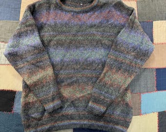 May include: A long-sleeved crew neck sweater featuring a fuzzy, textured knit pattern. The garment displays a variegated color palette of charcoal gray, deep purple, muted green, and hints of rust orange, arranged in horizontal bands throughout.
