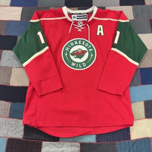 May include: A red Reebok Minnesota Wild hockey jersey featuring green sleeves with white accents, a white lace-up collar, the number 1 on each sleeve, and a large circular team logo on the chest with the letter A patch on the upper right side.