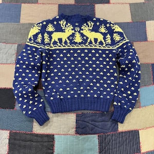 May include: A long-sleeve knit sweater in deep navy blue featuring a contrasting bright yellow pattern. The upper chest displays two reindeer facing a central pine tree, while the body and sleeves are covered in a repeating small dot motif.