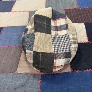 May include: A flat cap featuring a patchwork design composed of various fabric squares. The patterns include beige herringbone, black and white houndstooth, and dark gray plaid with thin red lines. The cap has a rounded crown and a short, stiff brim.