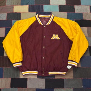 May include: A maroon and gold varsity jacket with the letter "M" on the chest. The jacket has gold sleeves, a maroon body, and white and gold striped trim on the cuffs, collar, and hem. The jacket is buttoned up.
