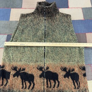 May include: A textured, zip-up vest in shades of green and brown, featuring a high collar and a black zipper. The bottom of the vest has a brown band with black moose silhouettes. The vest measures approximately 24 inches across.