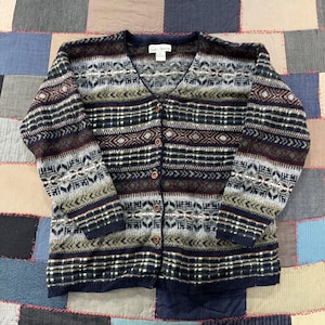 May include: A long-sleeve V-neck cardigan sweater featuring a multi-colored geometric and fair isle pattern in shades of navy blue, dark green, burgundy, and grey. The front includes five brown buttons and a label reading Croft & Barrow.