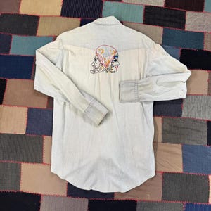 May include: Light blue denim shirt with long sleeves. The back features an embroidered design of two faces wearing a headscarf with a crescent moon and stars. The shirt has a button-down front and cuffs.