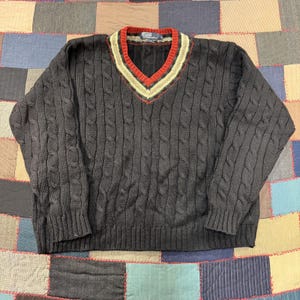 May include: A black cable-knit V-neck sweater featuring a ribbed collar with red and cream-colored stripes. The interior neck label displays the text Polo by Ralph Lauren. The long-sleeved garment has a textured knit pattern throughout the body.