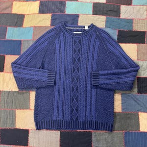 May include: A blue knit sweater with a textured cable-knit design. The sweater features a crew neck and ribbed cuffs and hem. The color is a deep shade of blue, with a lighter blue accent in the cable knit pattern.