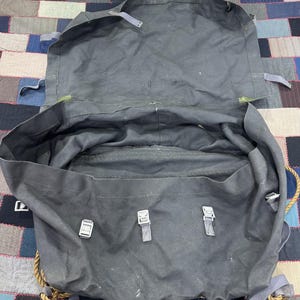 May include: A dark gray canvas rucksack with a large main compartment and a wide top flap. The bag features three metal buckles on the front, adjustable gray fabric straps, and thick tan rope detailing along the sides and bottom edges.
