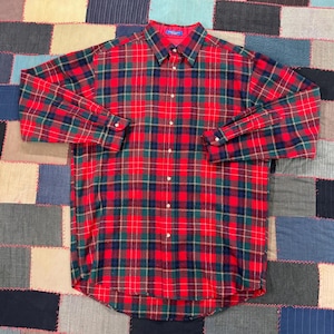 May include: A long-sleeved, button-down shirt in a classic plaid pattern. The shirt features a red base with green and navy blue intersecting lines, creating a traditional tartan design. The collar and cuffs are visible, and the shirt is buttoned up.