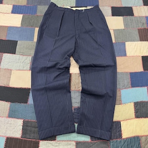 May include: Navy blue trousers featuring a white pinstripe pattern, a high-waisted design with double front pleats, a button and zipper fly closure, and straight-leg cuffs. The garment is constructed with a structured waistband and belt loops.