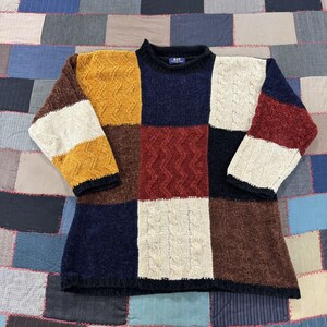 May include: A long-sleeve sweater featuring a multi-colored patchwork design with blocks of mustard yellow, navy blue, cream, dark brown, and deep red. The knit pattern alternates between vertical cable knit textures and smooth knit sections.