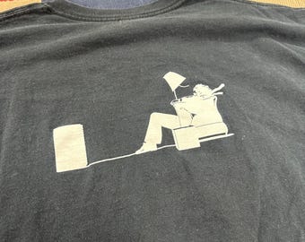 Pode incluir: T-shirt preto de gola redonda com um gráfico branco. O gráfico mostra uma pessoa sentada numa cadeira com uma lâmpada, uma mala e uma forma retangular. O design é simples e moderno.