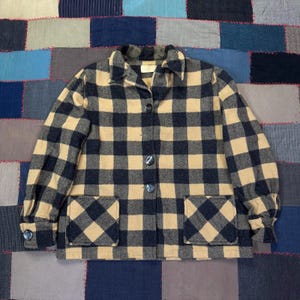 May include: A vintage, button-down jacket in a classic buffalo plaid pattern. The jacket features a tan and navy blue checkered design, two front pockets, and a collared neckline. The jacket has four buttons down the front and one on each cuff.