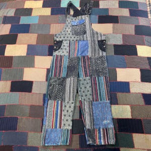 May include: Patchwork overalls in various shades of blue, gray, and black. The bib overalls feature a mix of geometric and striped patterns. The straps and pockets are a solid dark blue. The garment is displayed on a patchwork quilt.