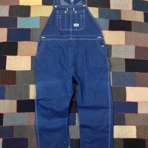 May include: Blue denim overalls with adjustable straps, a bib pocket, and side pockets. The overalls have white stitching and brass-colored hardware. The brand name is visible on the bib pocket. The background is a patchwork quilt.