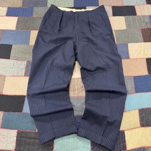 May include: Navy blue trousers featuring a white pinstripe pattern, a pleated front design, and a light-colored interior waistband lining. These pants have a straight-leg cut with cuffed hems and a button closure at the top of the waistband.
