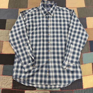 May include: A long-sleeved button-down shirt in a blue and white plaid pattern. The shirt has a classic collar and buttoned cuffs. The shirt is made of a medium-weight fabric and has a relaxed fit.