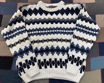 May include: A long-sleeve crew neck sweater featuring a repeating geometric pattern in shades of cream, navy blue, light blue, and olive green. The knit garment has a ribbed collar, cuffs, and hem, with a textured surface throughout the body.