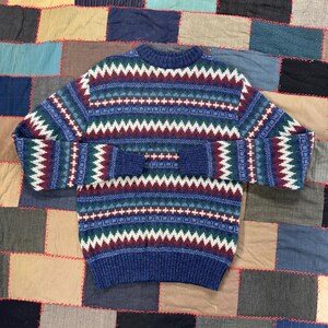 May include: A blue knit sweater with a crew neck and long sleeves. The sweater features a repeating pattern of white, green, and maroon zigzags and crosses. The sweater is displayed on a patchwork quilt.