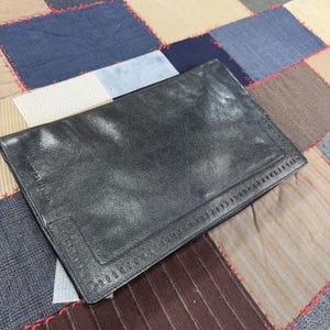 May include: A black leather clutch with a rectangular shape. The clutch has a stitched border and a flap closure. The background is a patchwork of various fabrics in shades of blue, brown, and gray.