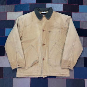 May include: A tan work jacket with a dark green collar. The jacket has four front pockets, button closures, and a small pocket on the chest. The jacket is made of a durable material.