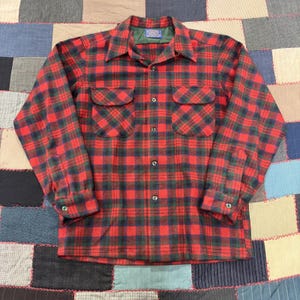 May include: A long-sleeve button-up shirt featuring a red, dark green, and navy blue plaid pattern. The shirt includes a pointed collar, two chest pockets with rounded flaps, and buttons on the front placket and cuffs. A Pendleton label is visible.