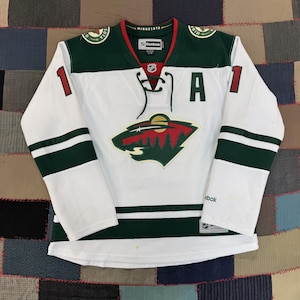May include: A white Reebok Minnesota Wild hockey jersey featuring a green and red team logo on the chest, a lace-up collar, the letter A on the left chest, and the number 11 on the sleeves. The jersey includes green and red accents and NHL branding.