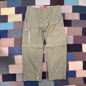 May include: Olive green convertible pants with multiple pockets and a zip-off design. Features a light-colored zipper and a button closure at the waist. The pants are made of a lightweight material.