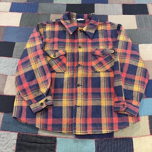May include: A long-sleeved, button-down flannel shirt in a classic plaid pattern. The shirt features a color palette of navy blue, red, yellow, and orange. It has two chest pockets and buttoned cuffs. The shirt is laid flat.