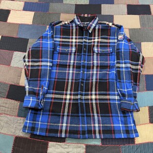 May include: A long-sleeve button-down shirt featuring a blue, black, and white plaid pattern. The shirt includes two chest pockets with button flaps, a standard collar, and a small Polo Sport logo patch on the upper left sleeve near the shoulder.
