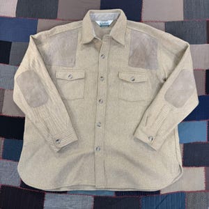 May include: A beige long-sleeve button-down shirt featuring a textured fabric, two chest pockets with button closures, and light brown suede patches on the shoulders and elbows. The shirt has a standard collar and a buttoned front placket.