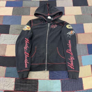May include: Black zip-up hoodie with red stitching and Harley-Davidson branding. Features a hood, front pockets, and patches on the sleeves. The Harley-Davidson logo is displayed on the chest and sleeves. The sleeves have the words "Harley-Davidson" in red.
