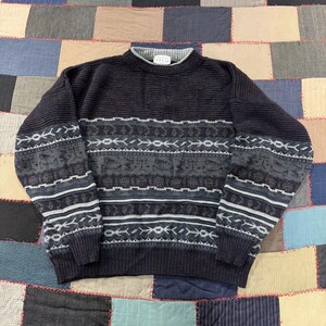 May include: A long-sleeve crew neck sweater in dark charcoal gray and black. The upper chest features a solid ribbed knit, while the lower body and sleeves display a horizontal pattern of geometric shapes and lines in shades of gray and white.