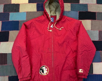 May include: A red hooded Starter jacket with a full-length zip. The jacket has a Seminoles logo patch on the left chest and a Starter logo on the right sleeve. The hood has a tan lining and drawstrings. The jacket is made of a lightweight material.