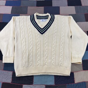 May include: A cream-colored long-sleeve V-neck sweater featuring a cable knit pattern on the front. The neckline is accented with dark green and navy blue stripes. The sweater has ribbed cuffs and a ribbed hem, with a small white brand label.